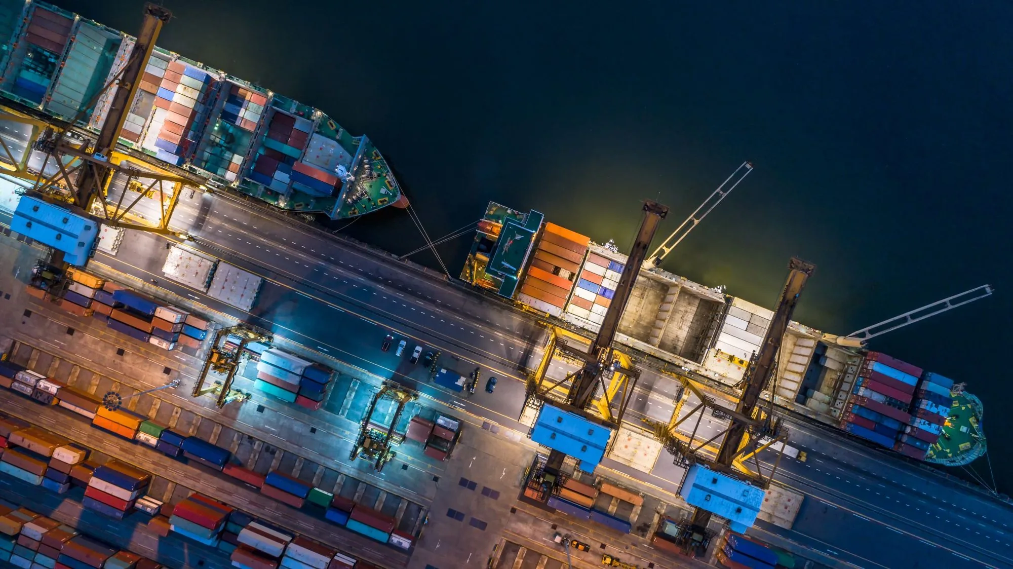 A floodlit container terminal at night seen from the air, with quay cranes and stacked containers