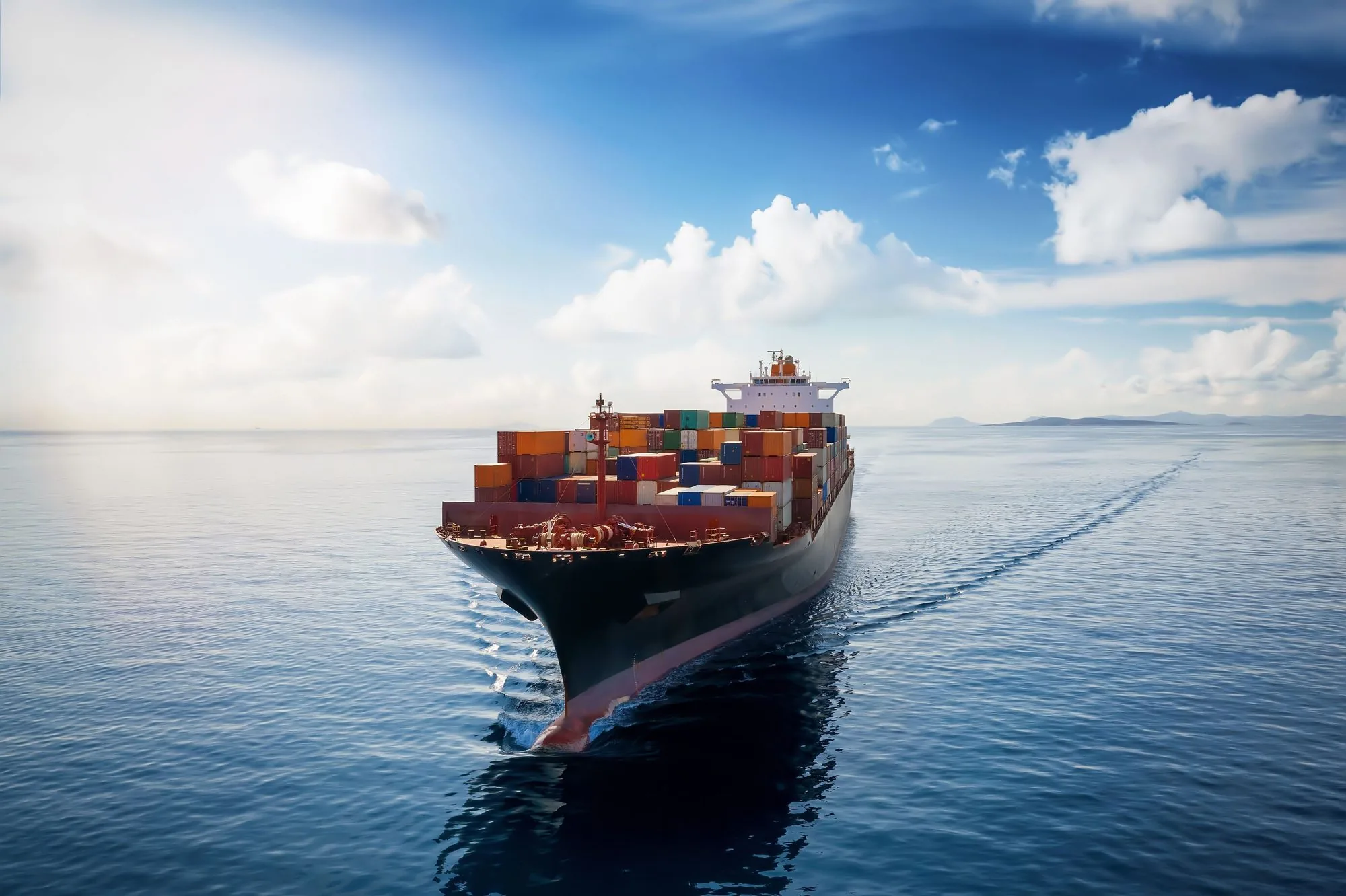 A container ship seen head on across a glassy sea under cumulus clouds