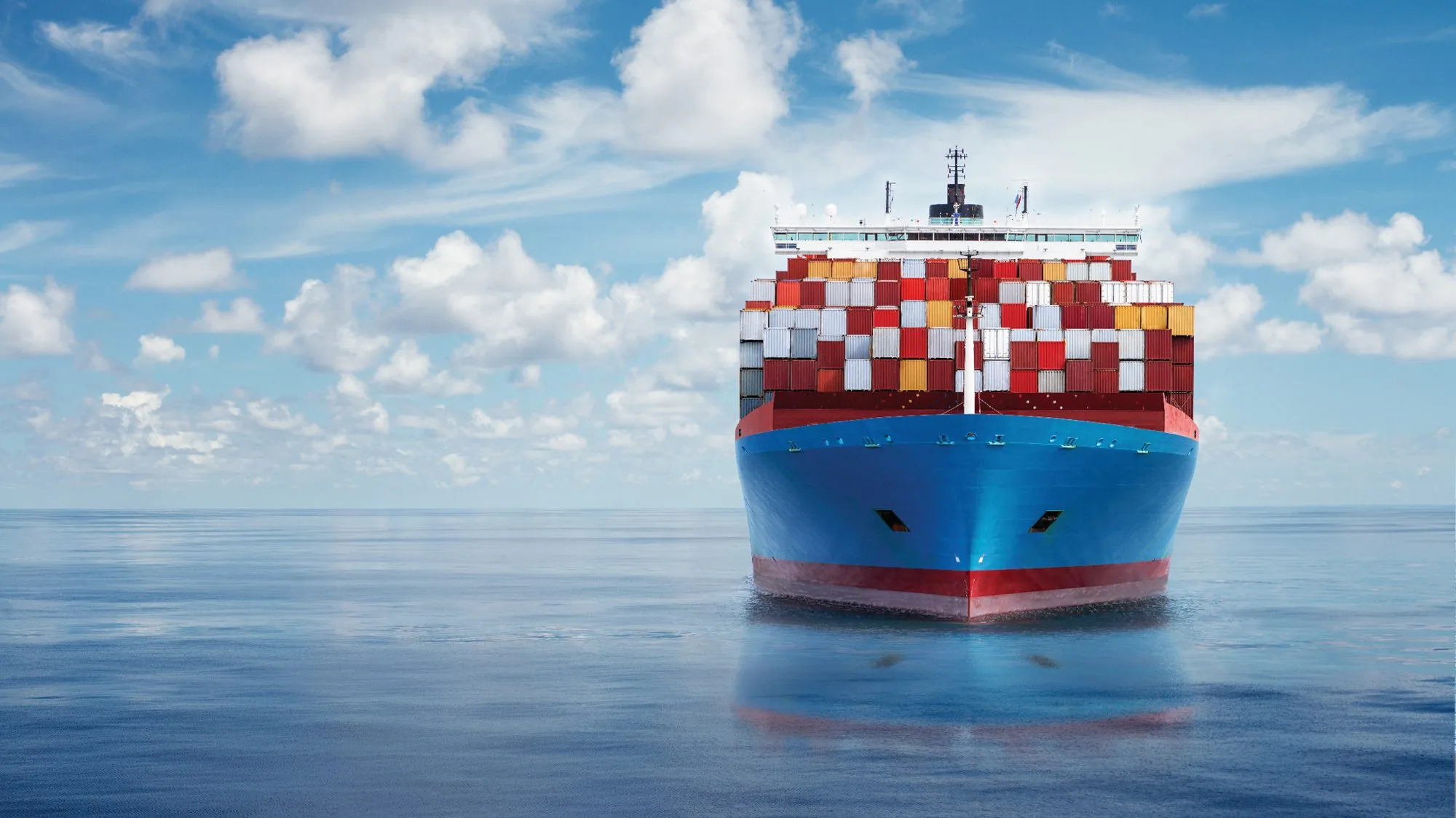 A blue hulled container ship seen head on across a mirror calm sea